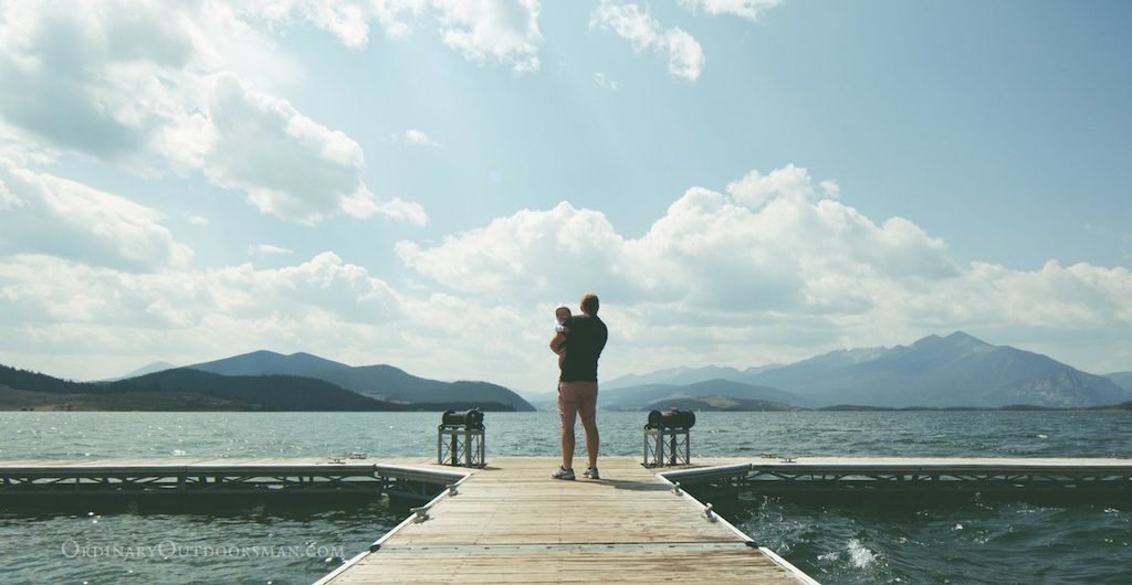 photo of a man holding an infant child while standing on a dock overlooking a mountain lake