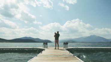 photo of a man holding an infant child while standing on a dock overlooking a mountain lake