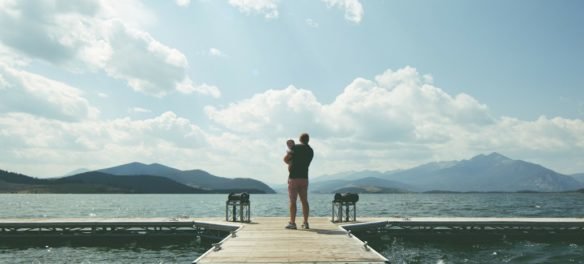 photo of a man holding an infant child while standing on a dock overlooking a mountain lake