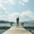 photo of a man holding an infant child while standing on a dock overlooking a mountain lake